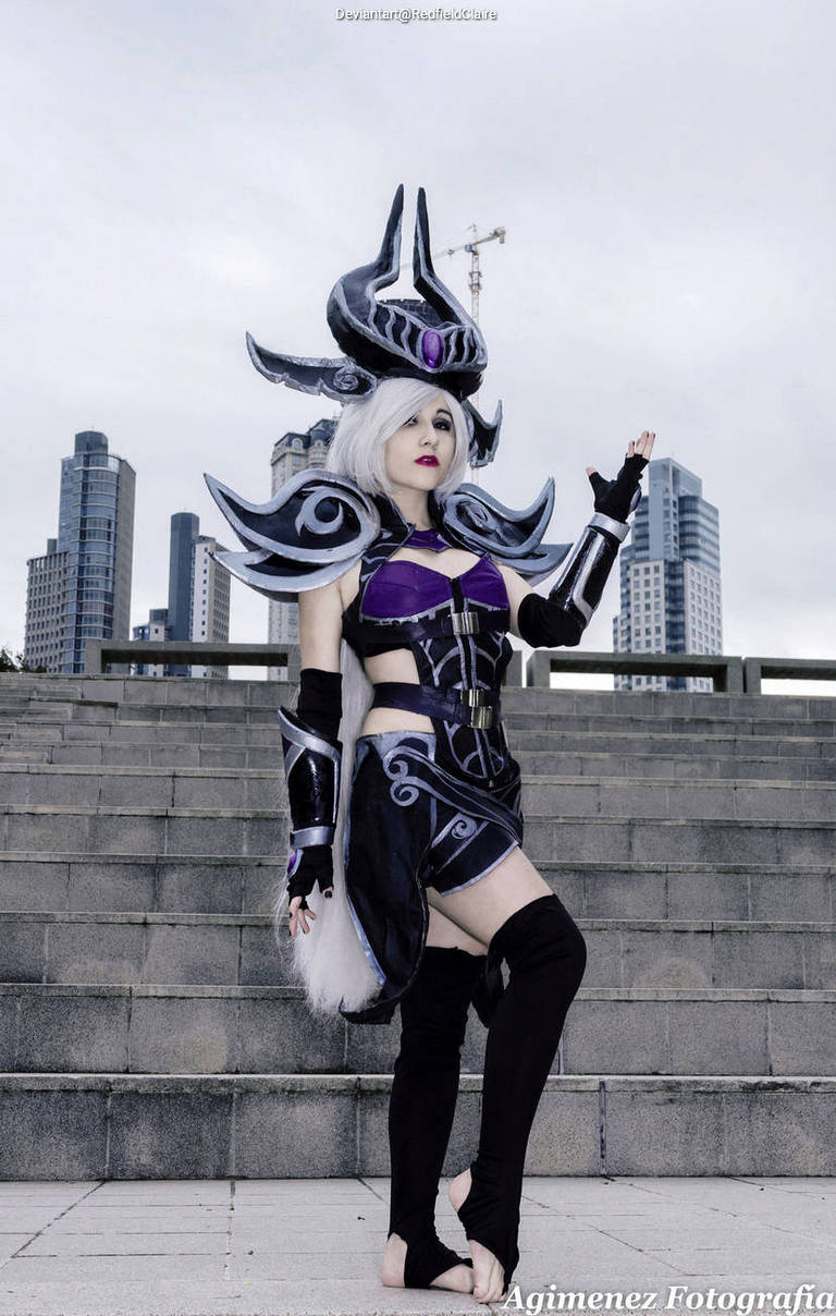 A person wearing an elaborate black and purple fantasy costume with armor-like details, striking a pose on outdoor steps with a city skyline in the background.