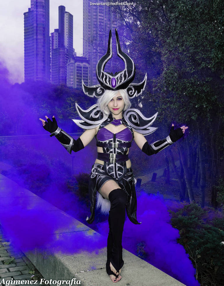 A person in an elaborate dark fantasy costume, featuring a horned headdress, standing confidently in an urban environment with purple smoke surrounding them.