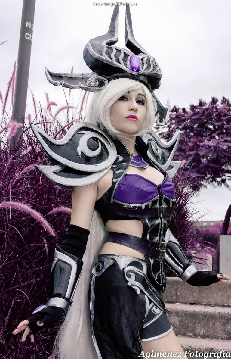 A person dressed in elaborate dark fantasy cosplay, featuring a intricate helmet with a purple gem, silver armor, and long white hair, poses confidently in a vibrant purple outdoor setting.