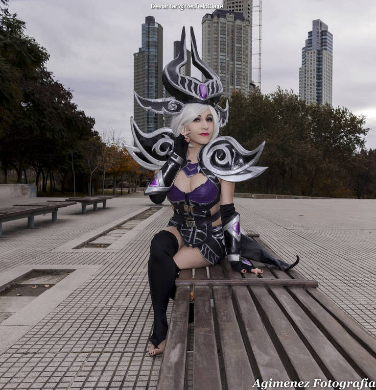 A cosplayer in a dramatic outfit with large shoulder armor sits on a bench in an urban setting, featuring tall buildings and autumn foliage in the background.