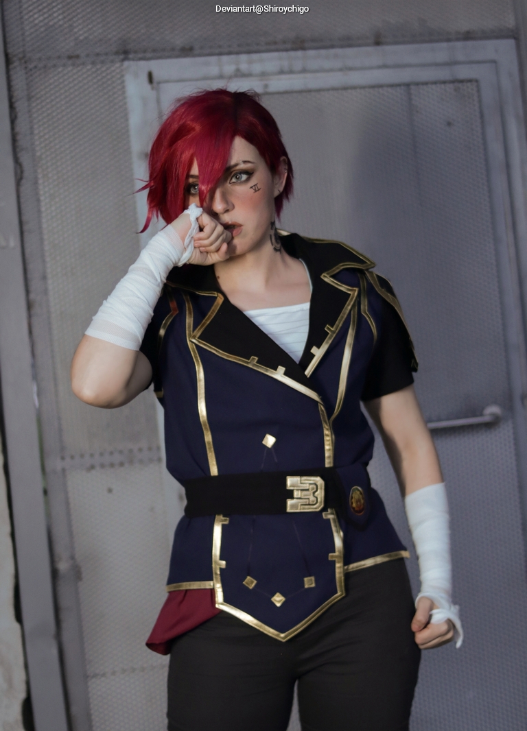 A person in a dark blue and gold cosplay outfit with short red hair, posing with one hand near their mouth and a serious expression.