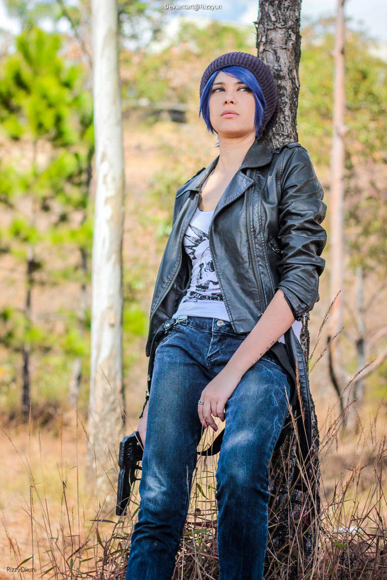 A person with blue hair wearing a black leather jacket and blue beanie is sitting against a tree in a natural outdoor setting, holding a toy gun.