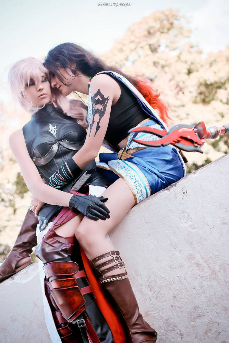 Two women in cosplay pose intimately, one wearing a black armor-like top and the other dressed in a colorful outfit with a sword. They are sitting closely together against a light-colored wall, with trees in the background.