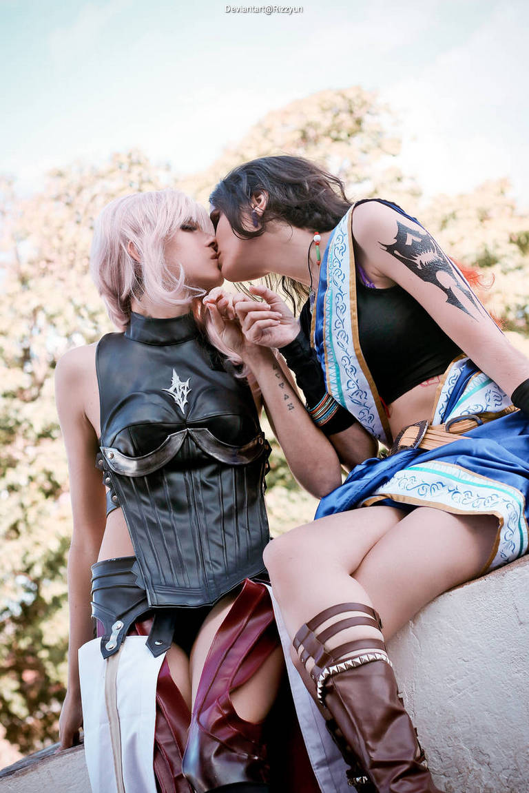 Two cosplayers dressed in fantasy-inspired outfits share a kiss, surrounded by greenery in a bright outdoor setting.