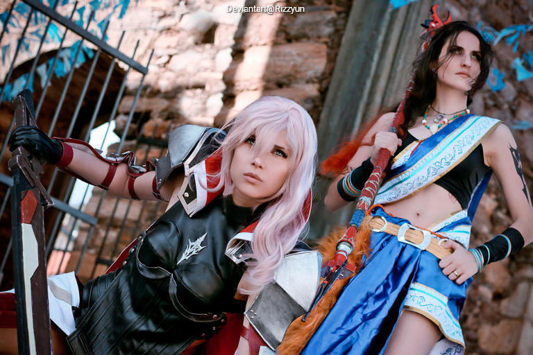 Two cosplayers posing in an outdoor setting. One wears a black and silver armored outfit with pink hair, while the other is dressed in a blue traditional costume with long dark hair, holding a staff.
