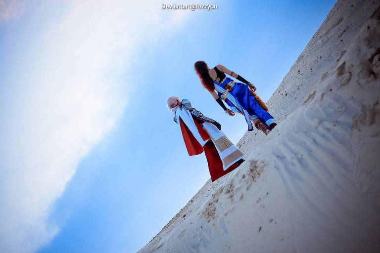 Two individuals dressed in elaborate fantasy costumes stand on a sandy dune, looking out towards a blue sky.