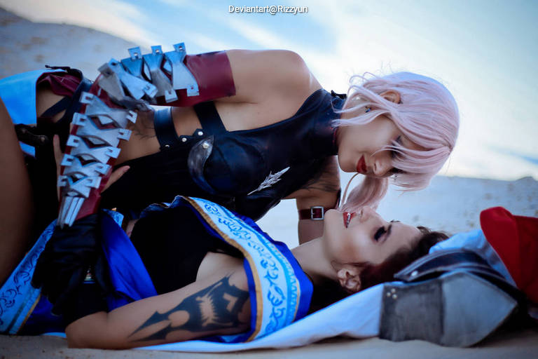 Two individuals in a playful pose on a sandy surface, one is lying down while the other leans over them. They are dressed in detailed costumes, showcasing vibrant colors and intricate designs.