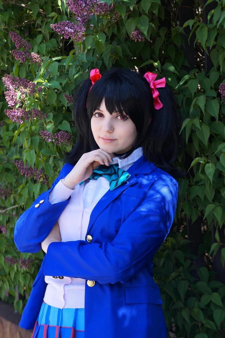 Young woman in a blue blazer and plaid skirt, standing against a backdrop of greenery and lilac flowers, with black hair styled in pigtails adorned with pink bows, posing thoughtfully.