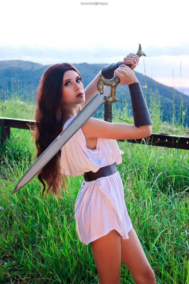 A woman in a white tunic with a black belt poses dramatically in a grassy landscape, holding a large sword with both hands, looking confidently into the distance.