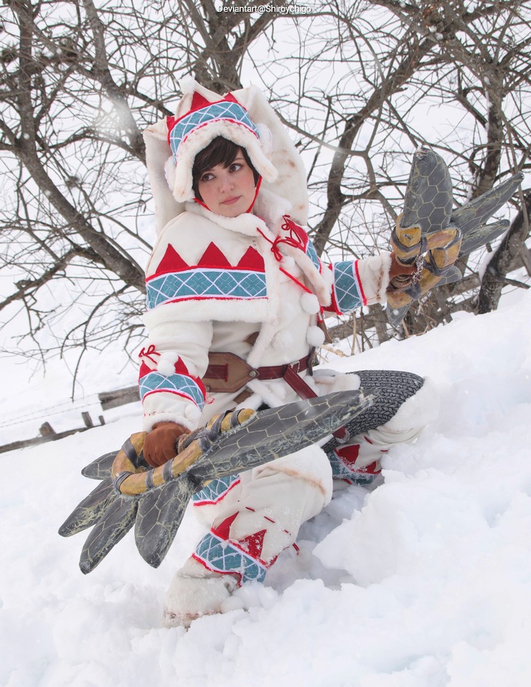 A person in a winter costume inspired by a traditional Inuit design, holding a large weapon resembling a fish, surrounded by snow and trees.