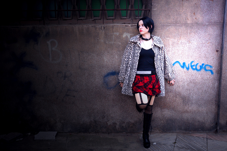 A young woman wearing a leopard print coat, a black top, and a red plaid skirt, standing against a wall with graffiti.