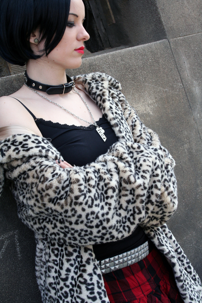 A young woman with a bob haircut, wearing a leopard print faux fur coat, black tank top, and plaid skirt, posing against a textured wall. She has red lipstick and a choker necklace.