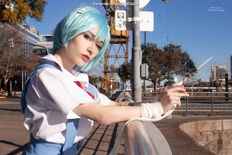 A person with blue hair dressed in a white and blue outfit, leaning against a railing in an urban setting, with cranes and buildings in the background.