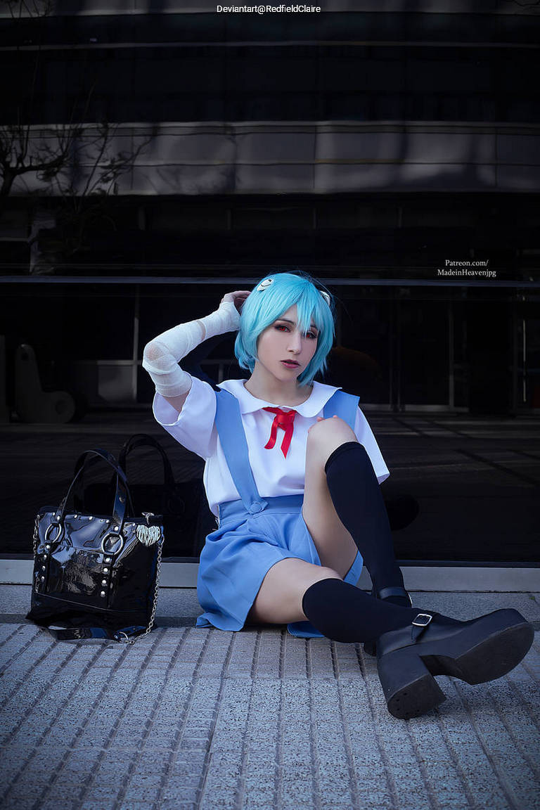 A person in a blue wig and outfit is sitting on a textured surface, wearing a white shirt with a red bow, a blue skirt, and black knee-high socks. They have a black handbag beside them.