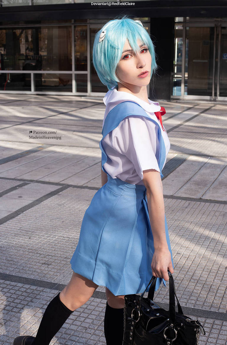 A person wearing a blue school uniform-style outfit with a white shirt and red bow, holding a black handbag. They have light blue hair and striking red eyes, posing confidently outdoors.