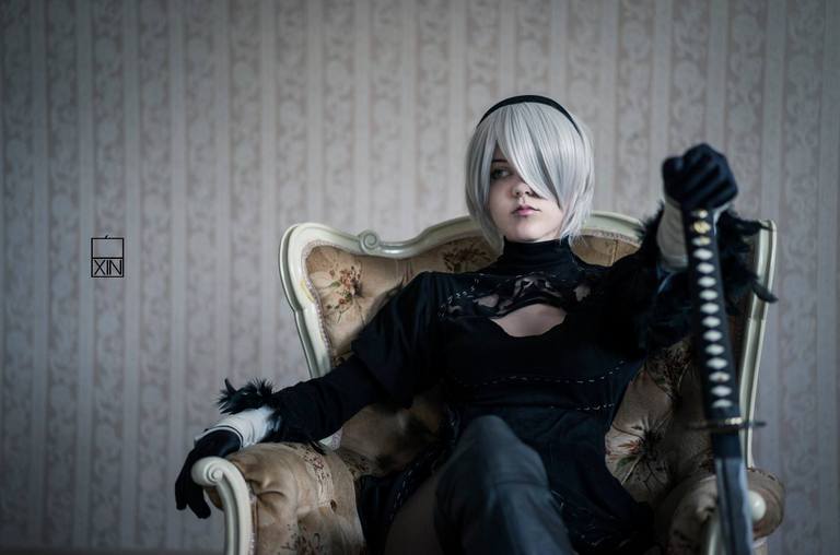 A person in a black costume with silver hair sits confidently in an ornate chair, holding a sword. The background features a patterned wall.