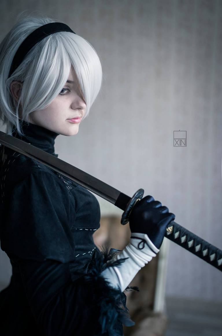 A person with short, platinum blonde hair wearing a black costume, holding a sword. The background is softly blurred, enhancing the focus on the character.