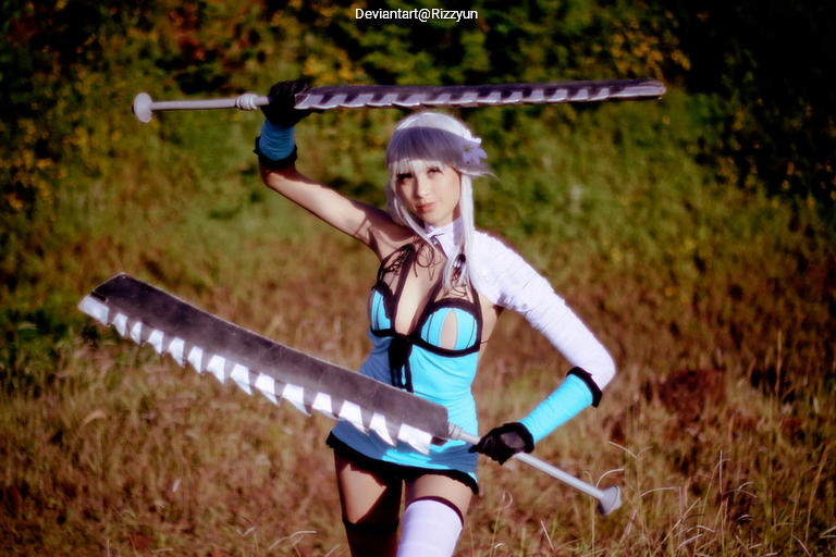 A cosplayer in a blue and black outfit holds two large swords, posing in an outdoor setting with greenery in the background.