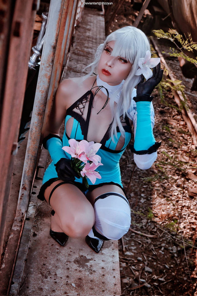 A woman in a blue costume with a black lace-up design, kneeling amidst a rustic background, holding pink flowers and wearing a white flower in her hair.
