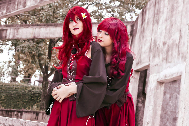 Two individuals dressed in matching red and black medieval-style outfits, posing together in an outdoor setting with greenery and a stone background.