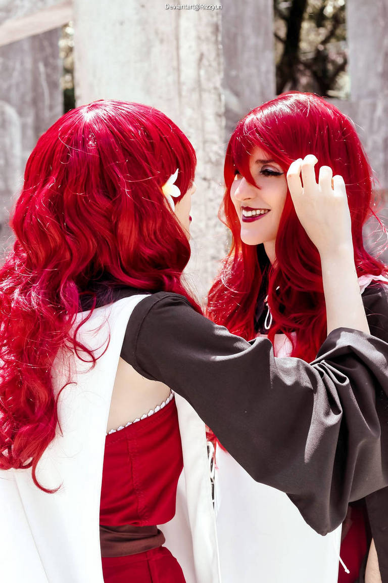 Two people with long red hair are interacting, one gently touching the other's hair. They are wearing costumes with a mix of dark and light colors, against a softly blurred outdoor background.