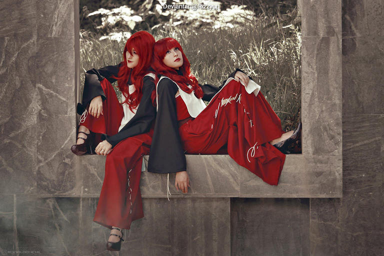 Two individuals with long red hair are seated against a stone structure, wearing matching red and black outfits. The background features greenery, creating a serene atmosphere.