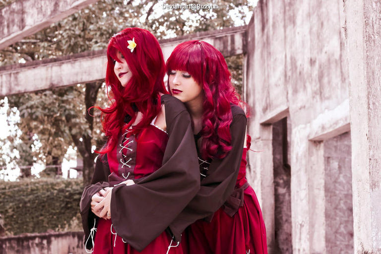 Two women with long red hair, wearing matching red dresses with black accents, are standing close together in a serene outdoor setting.