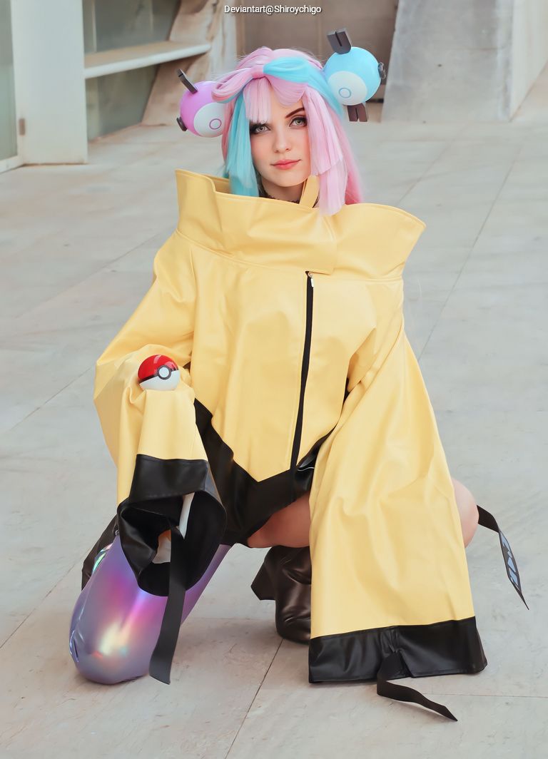 A person in a yellow oversized coat with black accents, kneeling on the ground. Their hair is styled with pink and blue colors, and they have playful accessories resembling headphones. The background features a modern architectural setting.