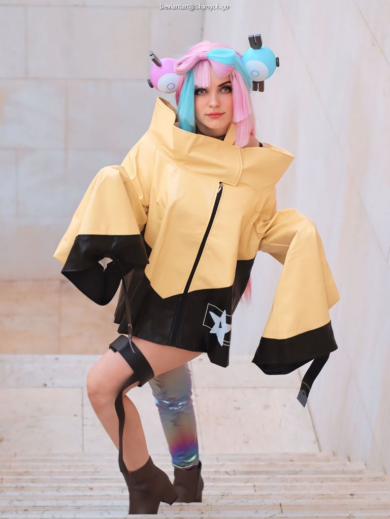 A person poses on stairs wearing a stylish, oversized yellow jacket with black accents and a star design. They have vibrant pink and blue hair styled in buns, complemented by shiny, colorful leggings and ankle boots.