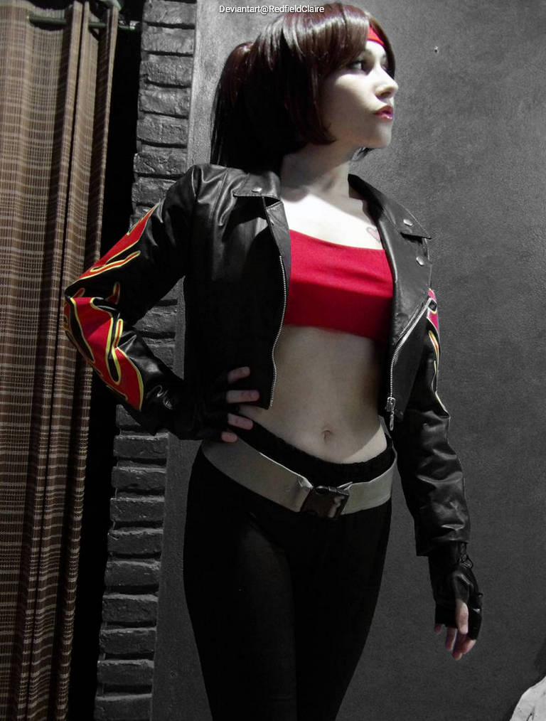 A person in a stylish black leather jacket with red and yellow details, wearing a red crop top and black pants, posing confidently against a textured wall.