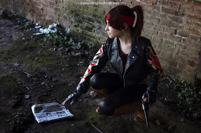 A cosplayer in a black leather jacket with red and yellow designs crouches down to examine a newspaper on the ground, holding a handgun in one hand, in an urban, gritty environment.