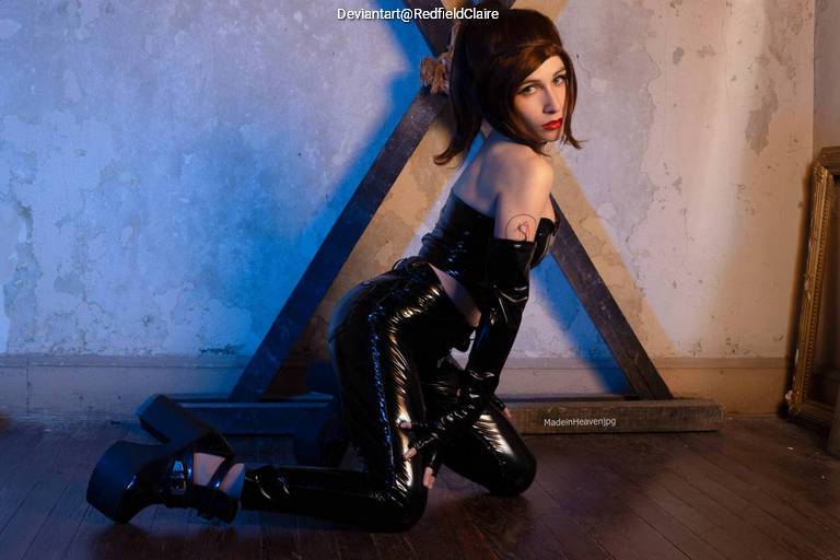 A woman in a shiny black outfit kneels on a wooden floor, posing against a textured wall with geometric wooden shapes behind her.