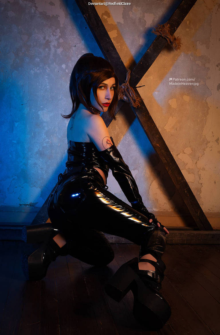 A figure in glossy black attire squats against a textured wall with wooden beams, illuminated by blue lighting.