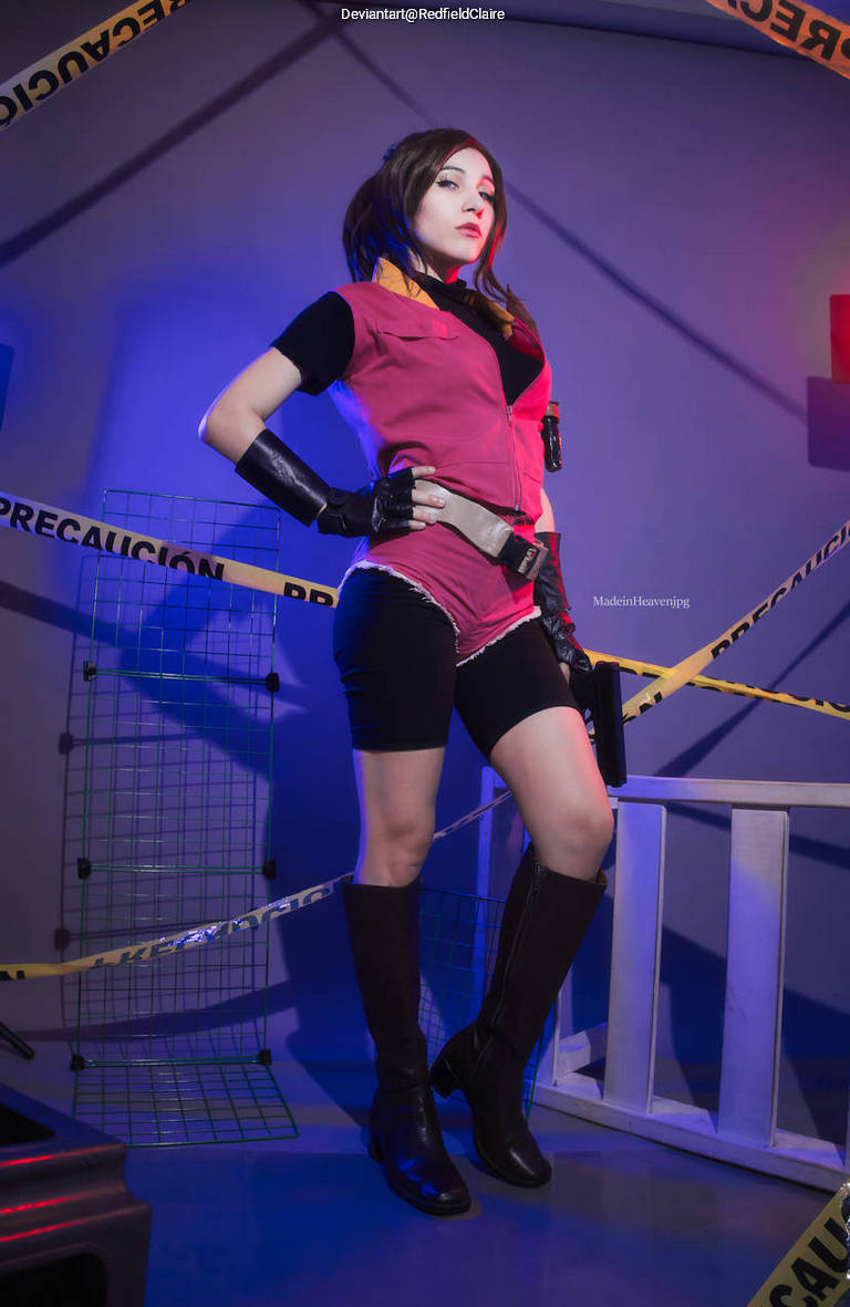 A woman cosplaying as a character with a red vest and black shorts, striking a confident pose against a backdrop with yellow caution tape and grid structures.