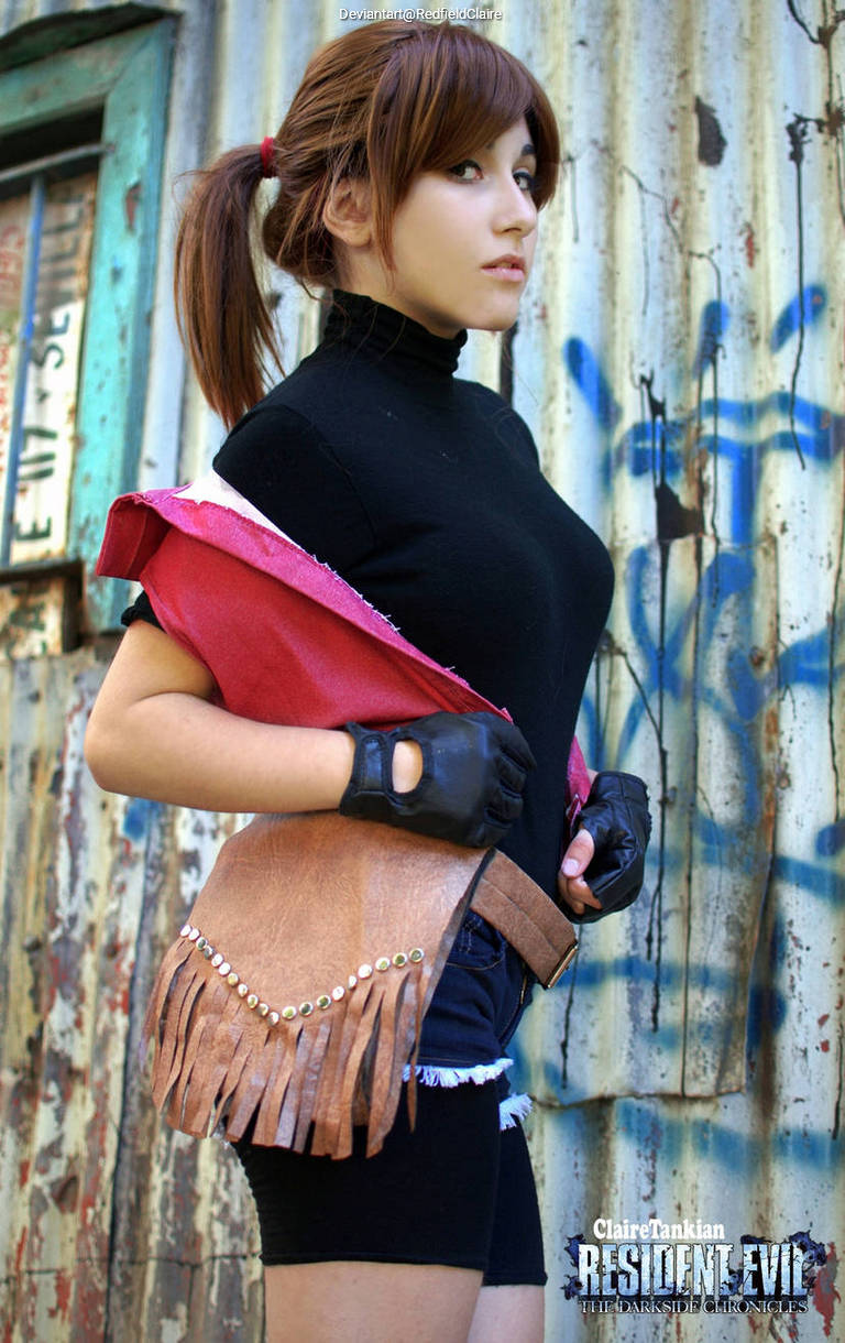 A woman in a black shirt and shorts poses in front of a peeling metal wall, wearing a red shawl and a fringed brown bag, with a serious expression.