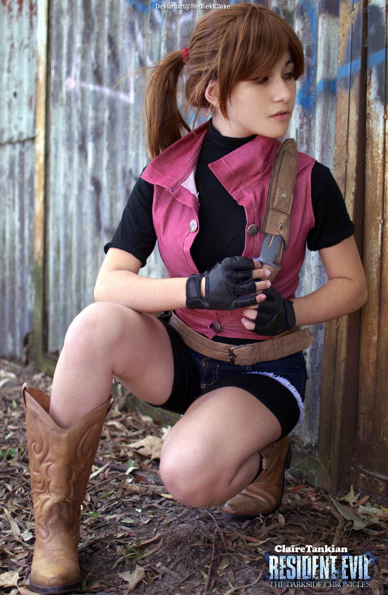A person in a pink vest and black shirt poses in a crouched position, wearing brown cowboy boots and gloves, against a weathered metal wall.