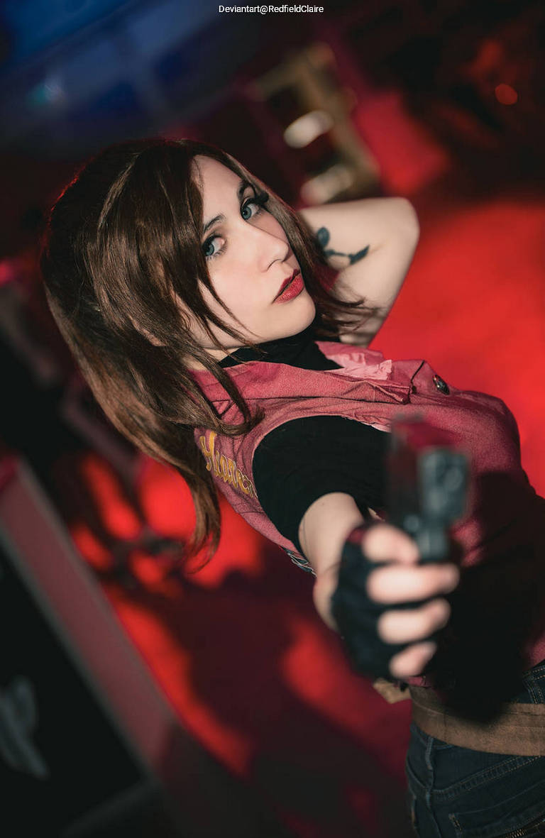 A woman in a pink vest points a handgun towards the camera, against a red-lit background.