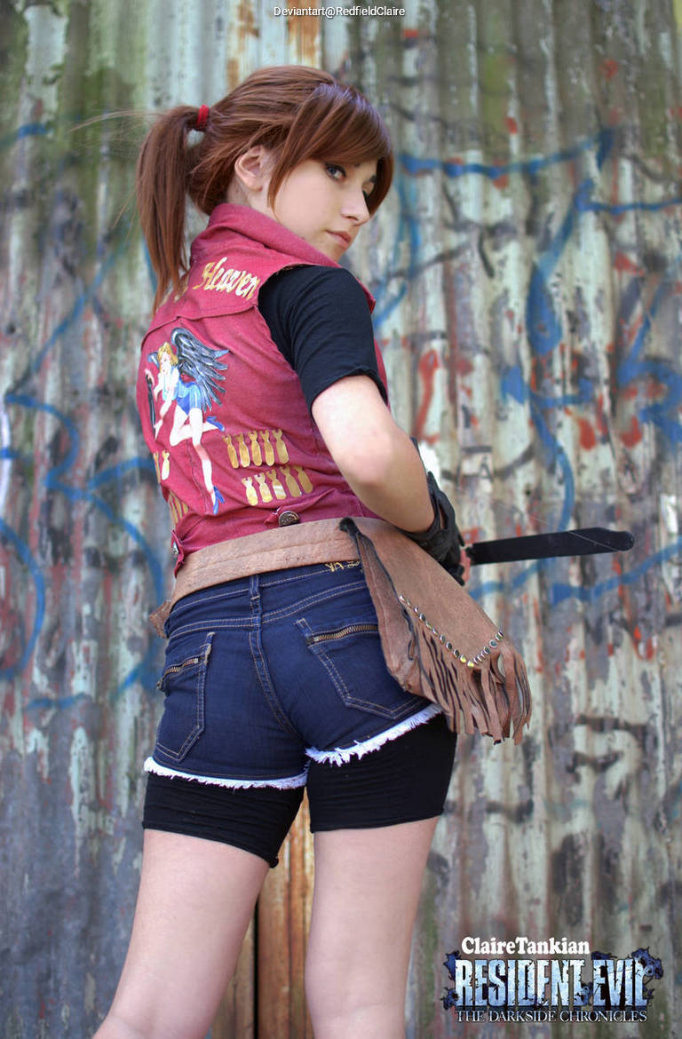 A person in a red vest with a graphic design, black shirt, and denim shorts stands against a graffiti-covered wall, showcasing a confident pose.