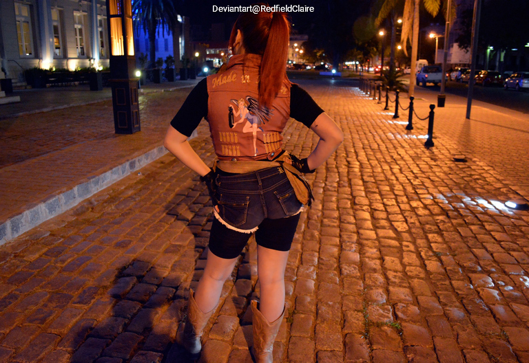 A person with long hair in a ponytail is standing on a cobblestone street at night, facing away from the camera. They are wearing a brown vest with a design on the back, a black shirt, and denim shorts, with cowboy boots. Streetlights illuminate the scene.