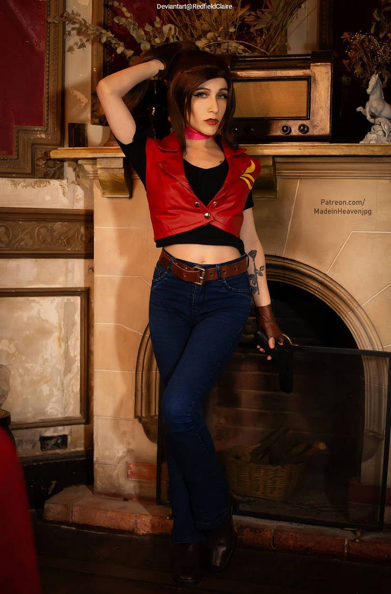 A person in a red vest and black shirt poses with one hand on their hip in a vintage-style room with a fireplace and old furniture.