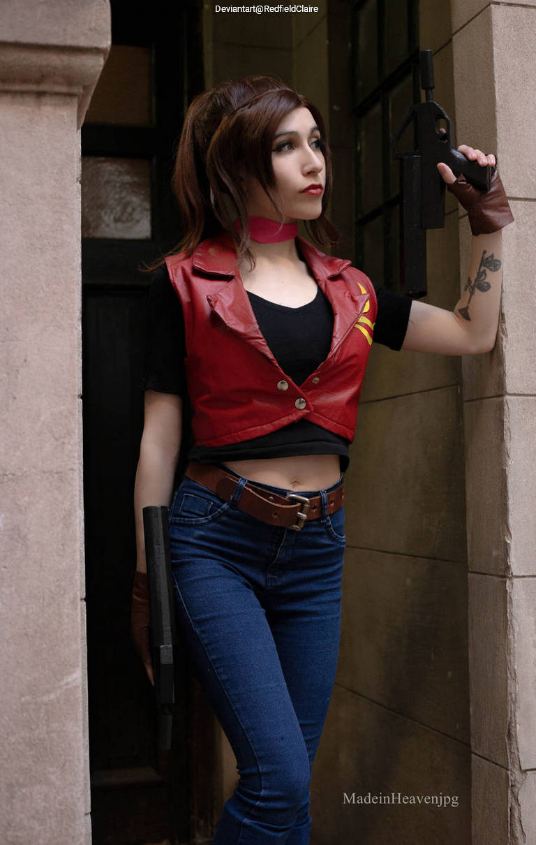 A person posing in a doorway, wearing a red leather vest and blue jeans. They are holding a weapon in one hand and have a confident expression.