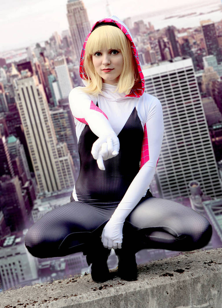 A female character in a stylized superhero costume, featuring a black bodysuit with pink accents and a hood. She is crouched on a city ledge, pointing forward, with a city skyline in the background.