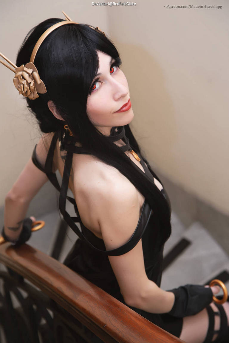 A cosplayer with long black hair and red eyes poses on a staircase, wearing a black outfit with straps and accessories, with a wooden railing in the background.