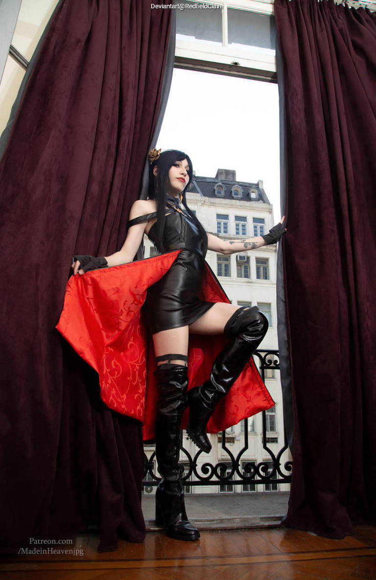 A person in a black leather outfit and thigh-high boots poses dramatically by a large window draped with dark curtains, holding a red and gold shawl.