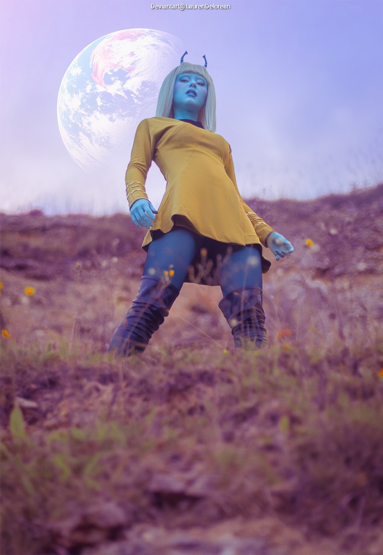 A blue-skinned character with horns stands on a hillside wearing a yellow dress, with a large planet visible in the sky behind them.