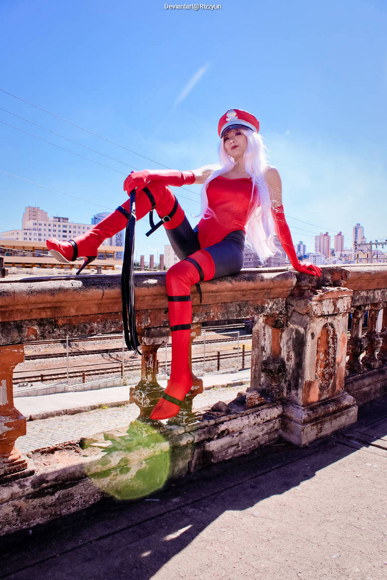 A person in a vibrant red and black costume is sitting on a stone railing with a city skyline in the background. The character has long white hair and is striking a confident pose, with one leg draped over the railing.