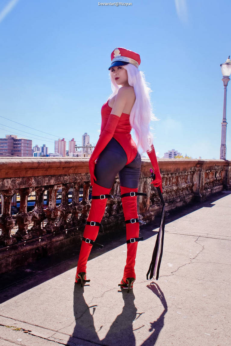 A cosplayer in a striking red outfit with long sleeves and a fitted bodice, wearing thigh-high red stockings and high heels. They have long white hair and a red cap, standing confidently on a sidewalk with a cityscape in the background.