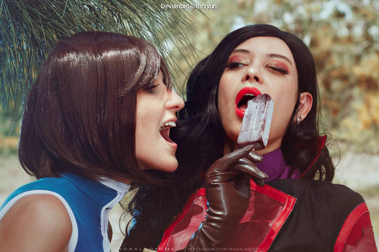 Two women in cosplay, one with short brown hair and a blue outfit, and the other with long black hair wearing a red and black costume. The woman in black is playfully eating a popsicle while the other looks on excitedly.