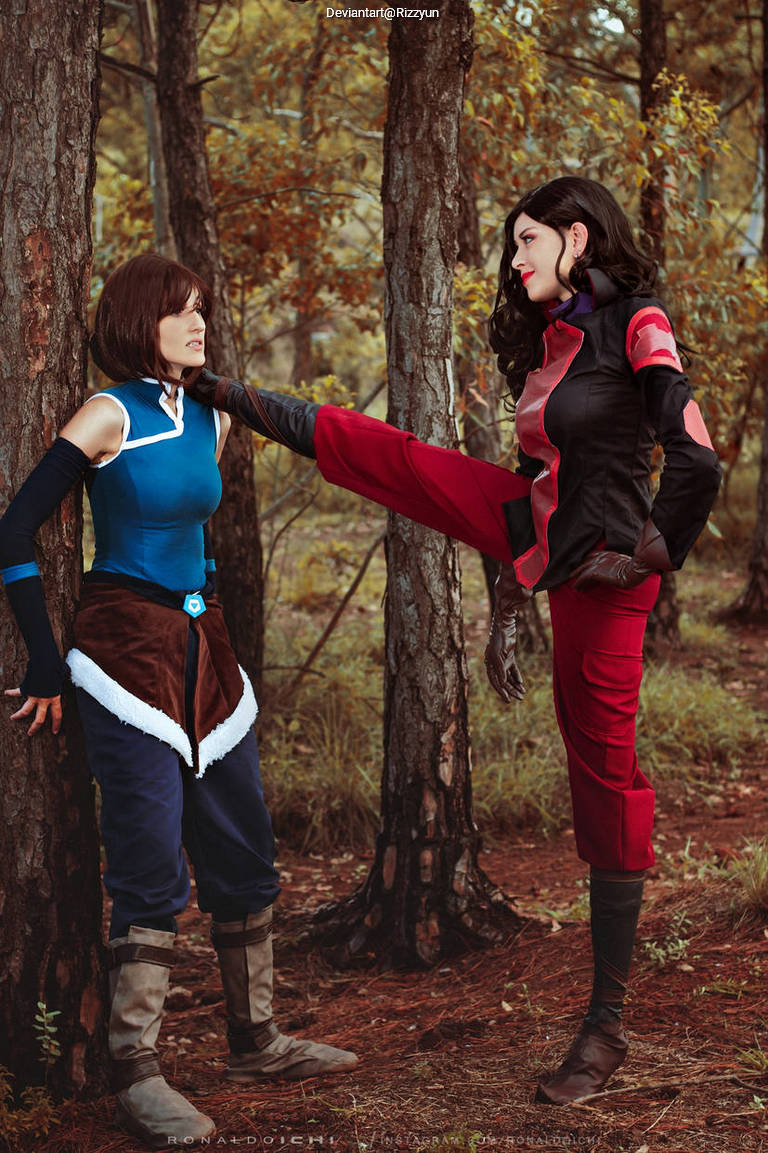 Two female cosplayers in a forest setting, one in blue and brown attire, the other in black and red, engaging in a dynamic standoff.