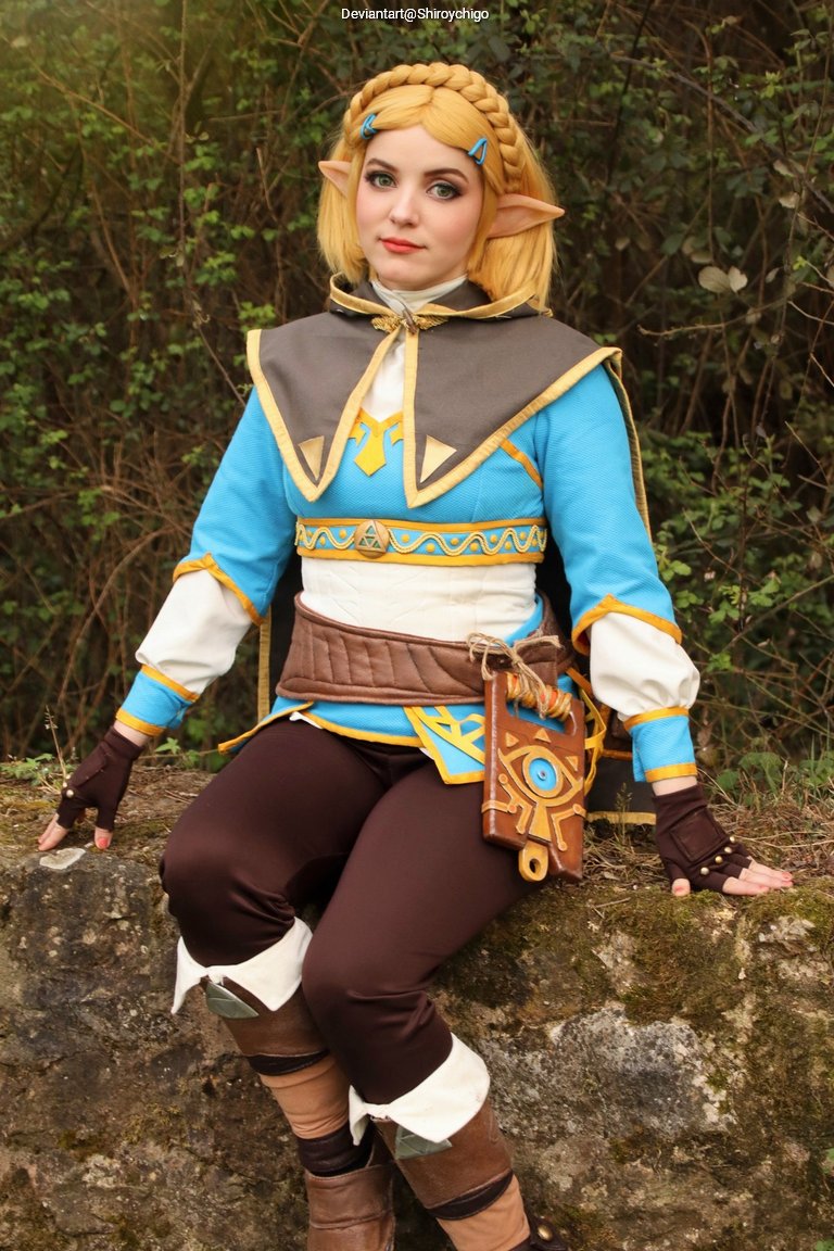 A person cosplaying as a character with blonde hair and pointed ears, wearing a blue and brown outfit with a decorative design, seated on a stone in a forested area.